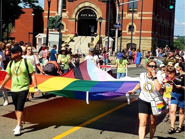 A group walks with an enormous Pride flag in front of Fredericton's city hall in the 2024 Fierté Fredericton Pride parade.