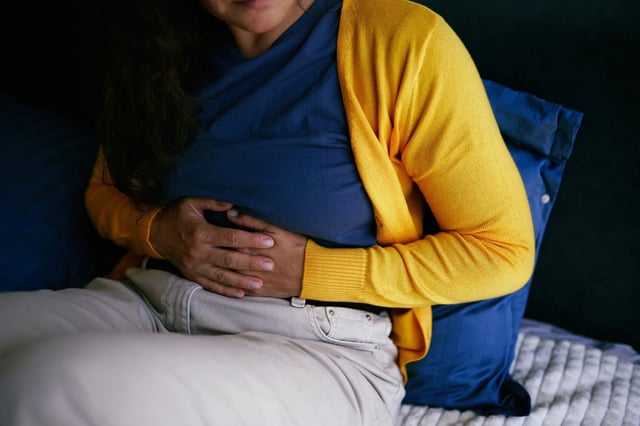 Close-up of a person seated and holding their stomach, depicting abdominal discomfort or pain. Yellow cardigan, blue shirt, and home setting elements provide a relatable and impactful representation.