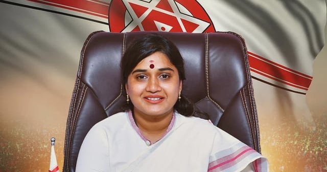 A woman dressed in a white saree with a pink border is seated at a wooden desk, hands clasped, smiling confidently. She has a red bindi on her forehead and is wearing gold bangles and earrings. Behind her is a large flag featuring the Janasena Party symbol—red and white stripes with a red circle and white star in the center. A small Janasena flag is placed on the desk to her left, and a neat stack of files is on her right. The background includes a large, cheering crowd, adding to the political tone of the image.