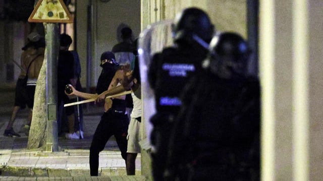 Eine vermummte Person hält einen Gegenstand wurfbereit, rechts vorne im Bild sind zwei Polizisten in Schutzausrüstung zu sehen