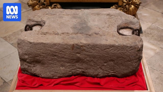 A large sandstone block sits on red carpet with two iron rings imbedded in it