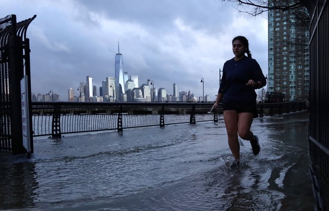 Water from the Hudson River can be seen overflowing a river wall at high tide as a woman jogs in front of the skyline of lower Manhattan in New York City on January 13, 2024, in Jersey City, New Jersey. (Photo by Gary Hershorn/Getty Images)