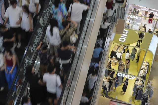 People take escalators as shoppers, at right downstairs, select clothing at a fashion boutique store inside a shopping mall, in Beijing on Sunday, July 13, 2025. (AP Photo/Andy Wong)