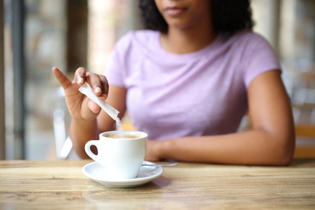 A stock image of a woman adding sugar in a coffee cup.