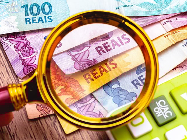 Real Brazilian currency. A man with magnifying glass looking at - BRL banknotes. green calculator in the composition. Time of financial crisis inflation and debts. FTP CLARIN Moneda de Brasil. Foto Shutterstock (2).jpg Z Invitado