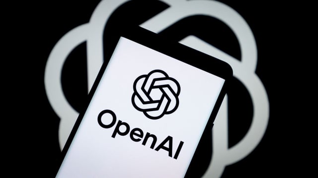 A smartphone screen shows the OpenAI logo in sharp focus, positioned in front of a larger, blurred version of the same logo in the background. The image visually represents the tech brand's expanding digital presence and product evolution.