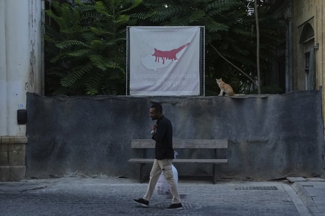 A man walks in front of a blocked road across the U.N buffer zone as a cat sits next of a banner shows the Cyprus island divided, the Turkish occupied area at the north and Cyprus republic at the south, in divided capital Nicosia, Cyprus, Thursday, July 17, 2025. (AP Photo/Petros Karadjias)