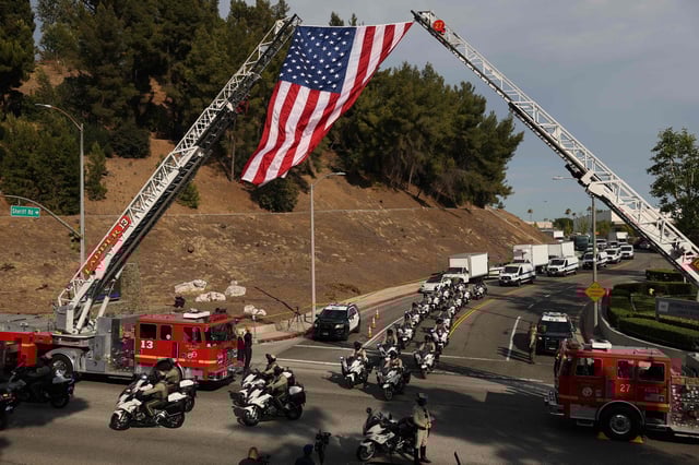Camiones de bomberos izan una bandera estadounidense mientras una procesión de camionetas del Servicio Forense con escoltas policiales y del sheriff pasa tras una explosión en el Centro de Entrenamiento Biscailuz del Departamento del Sheriff del Condado de Los Ángeles, en el Este de Los Ángeles, California, el 18 de julio de 2025