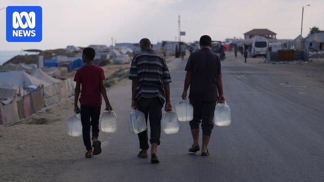 Three young men walk away from an aid distribution site, each carrying two heavy plastic bottles filled with water