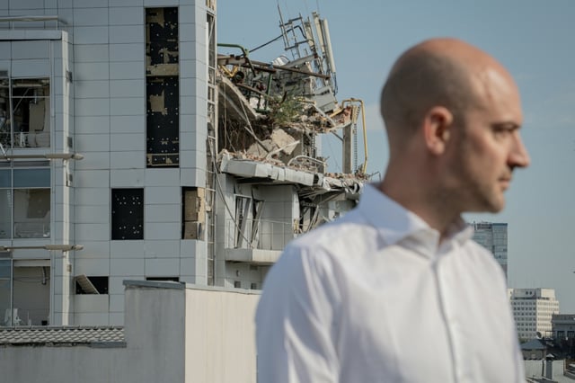Barrot is standing on a rooftop overlooking the city, from where ruined offices and residential buildings can be seen