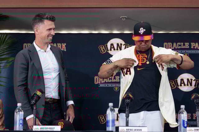 Buster Posey looks on as Rafael Devers puts on his New Jersey during a press conference at Oracle Park in San Francisco to announce that Devers has left the Boston Red Sox to join the San Francisco Giants on Tuesday June 17, 2025.