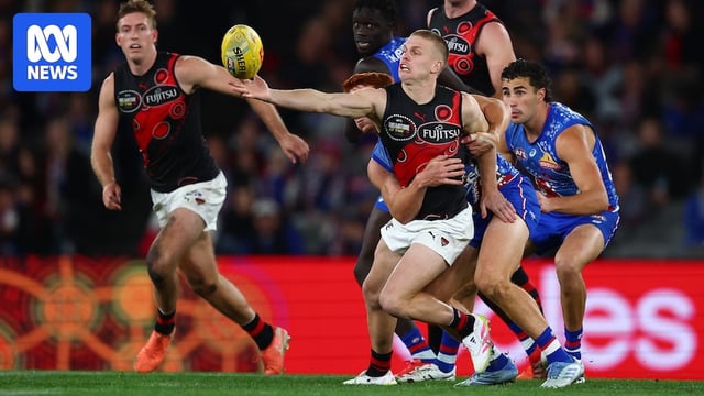 An Essendon AFL player reaches out to grab the ball as he is tackled from behind by a Western Bulldogs player.