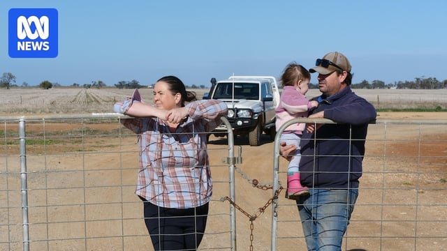 Woman leans on farm gate, man holds small child, white ute and paddock in the background.