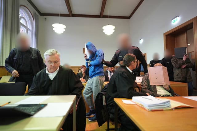 Die Angeklagten werden in Handschellen von Justizbeamten zu Beginn des Mordprozesses in den Sitzungssaal im Landgericht geführt.