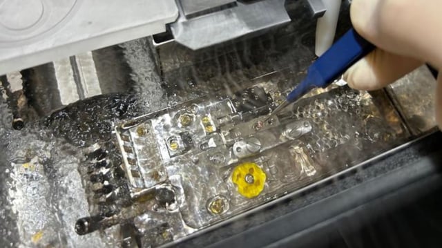 A gloved researcher uses precision tools to handle a sample on the cryo-TEM stage, surrounded by metallic components and cooling elements. The setup is submerged in liquid to maintain freezing conditions essential for high-resolution imaging of organic nanomaterials.