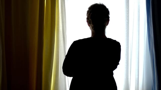 Eine Frau blickt im Gegenlicht aus einem Fenster mit Vorhang.
