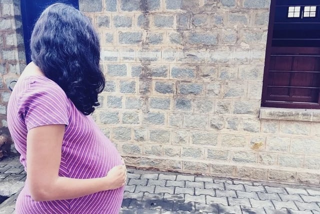A pregnant woman wearing a purple striped dress is standing sideways on a paved path, gently holding her belly with one hand. She has long, wavy black hair and is facing a stone wall with a window in the background.
