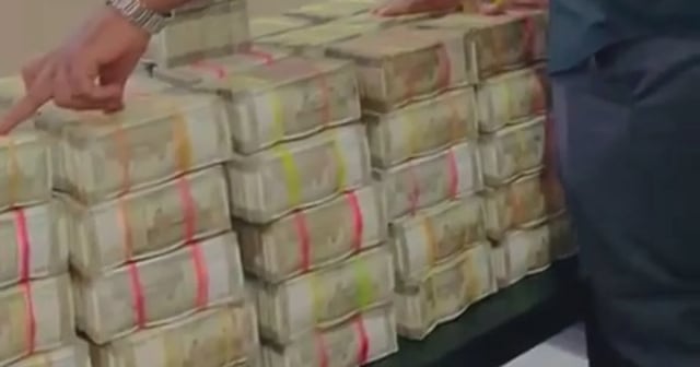 A man in a black shirt is standing beside a table stacked high with bundles of Indian currency notes, held together with colored bands. He appears to be pointing at one of the stacks, while resting his other hand on the money. The scene is indoors, with green and patterned curtains visible in the background.