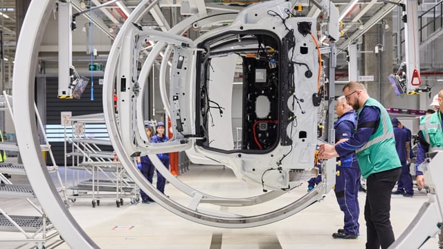 Workers look at a BMW chassis on a rotating jig at a factory.