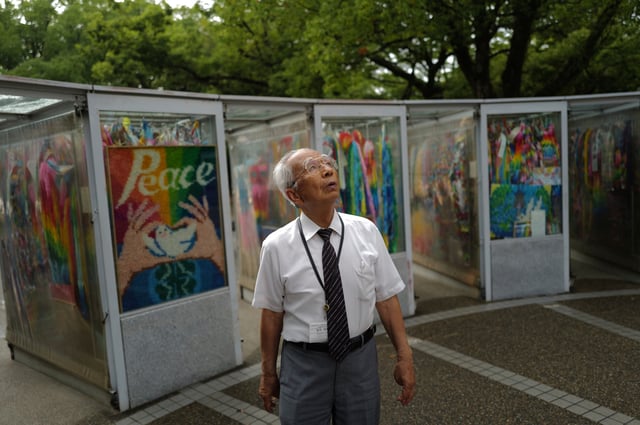 Kunihio Iida, atomic bomb survivor and a volunteer guide speaks in English to foreign visitors looks app the sky in front of the Children's Peace Monument where the place where people offer paper cranes to honor the victims of the 1945 atomic bomb on Wednesday, July 9, 2025, in Hiroshima, western Japan. (AP Photo/Eugene Hoshiko)