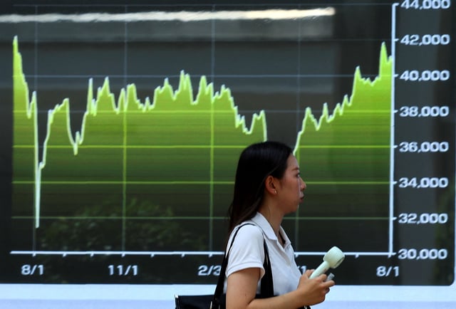 A woman holding a portable fan walks past a screen displaying a graph of recent Japan's Nikkei share average movements as the Japanese government issued a heatstroke alert in Tokyo and other prefectures due to a heatwave in Tokyo, Japan, August 4, 2025. REUTERS/Issei Kato/File Photo