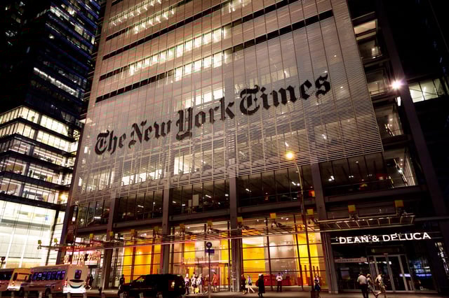 The headquarters of The New York Times company are seen at night. The glass-and-concrete building is behind a by a fronting composed of thin ceramic rods held horizontally, and at the lower floors the NYT's masthead logo is painted across them.