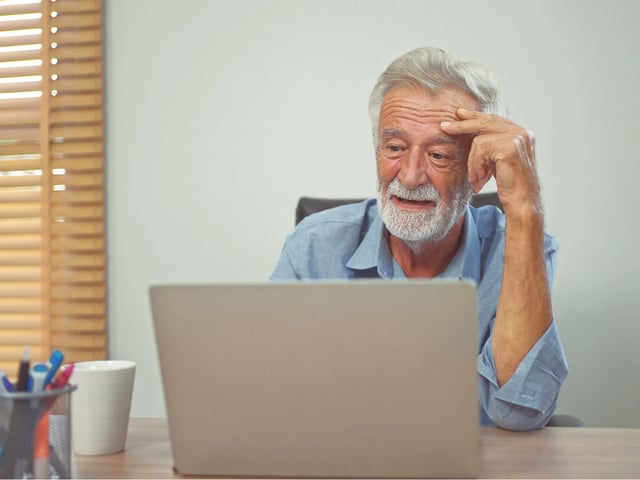 A worried older man uses a laptop at his kitchen table.