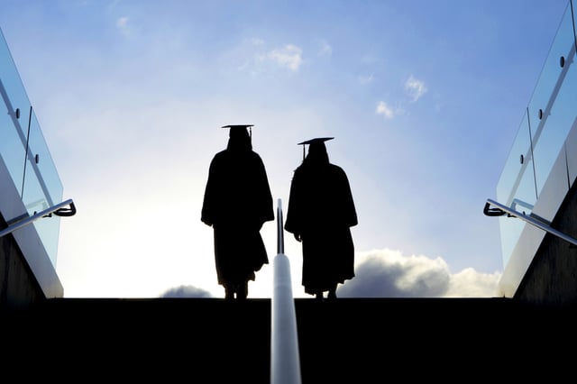 A shot of two college graduates as they make their way up a stairway and into the morning sunlight. They are dressed in graduation robes and hats. Lots of excitement and promise ahead of them.