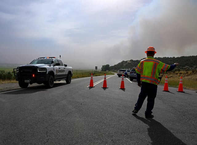 A Colorado Department of Transportation employee directs traffic at the intersection of W. Highway 64 and Rio Blanco County Rd. 7 as a Rio Blanco County Sheriff’s truck passes by while smoke billows from the Lee fire in Rio Blanco County just outside of Meeker on Thursday, Aug. 7, 2025. (Photo by Andy Cross/The Denver Post)