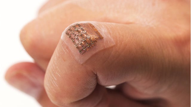 Representational image showing a small, transparent, flexible electronic patch adhered to the skin on a fingertip. The patch contains intricate metallic circuit patterns, highlighting the concept of wearable, skin-like electronics designed for continuous health monitoring.