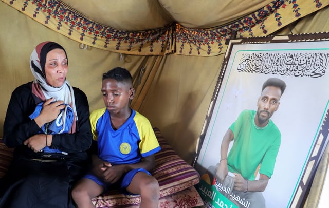 Doaa, the widow of late soccer player Suleiman Al-Obeid, known as the "Palestinian Pele," who was killed by an Israeli strike targeting people waiting for humanitarian aid, according to the Palestine Football Association, looks at his picture while holding his shorts, as her son sits beside her inside their tent in Gaza City August 9, 2025. REUTERS/Ebrahim Hajjaj