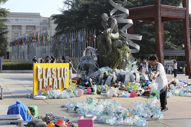 Benjamin Von Wong, right, a Canadian artist and activist, heaps piles of plastic waste onto a large sculpture that he designed in front of the United Nations office in Geneva, Switzerland, Monday, Aug. 11, 2025. (AP Photo/ Jennifer McDermott)
