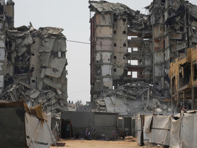A group of Palestinians sit in the shade of their tent amid destroyed buildings on a hot summer day in Gaza City, Tuesday, Aug. 12, 2025. (AP Photo/Jehad Alshrafi)