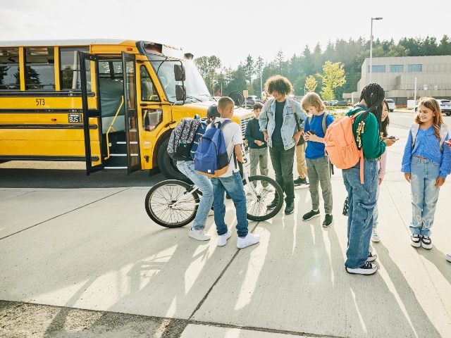Wide shot of middle school students hanging out and looking at phones after school waiting to board school bus for commute home