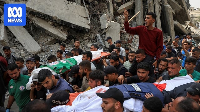 A crowd of men carry two covered bodies in stretchers past ruined buildings.