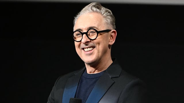 A man with glasses and a black suit is smiling