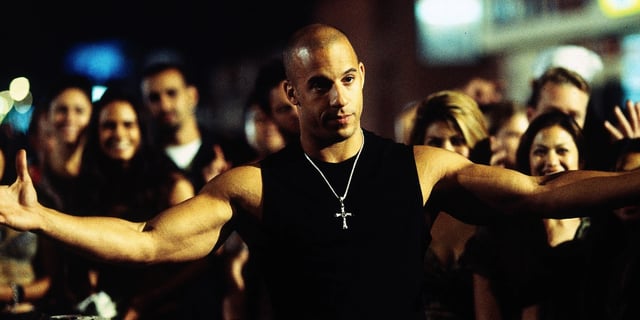 Vin Diesel as Dominic Toretto holding his arms out with a crowd behind in The Fast and the Furious, 2001. 