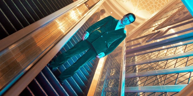 Colin Farrell wears a lavish suit and sports a thin mustache while riding down an escalator 