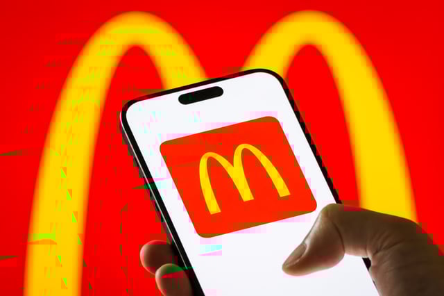 Photo illustration of a person holding a smartphone displaying the McDonald&#39;s logo against a background of the same logo.