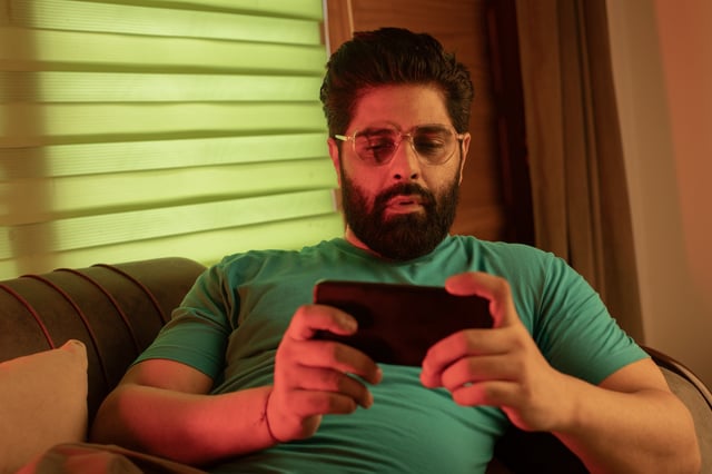 Young man in eyeglasses playing video game over smart phone while lying on sofa in living room