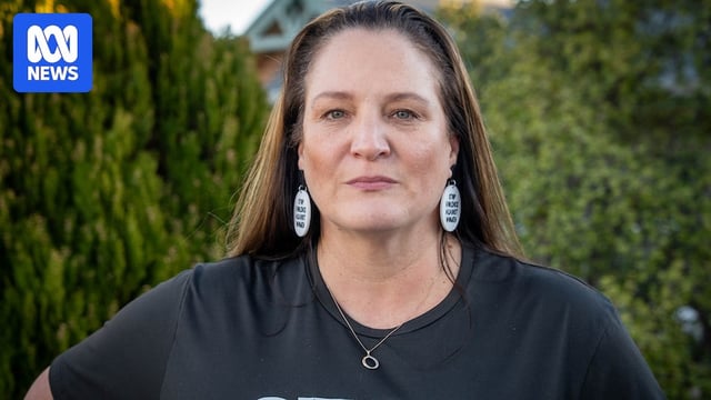 A woman has long brown hair, she looks solemn. She wears earrings which read "stop violence against women"