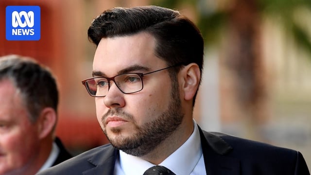 A young man with dark hair, a beard, a suit and glasses walks on the street.