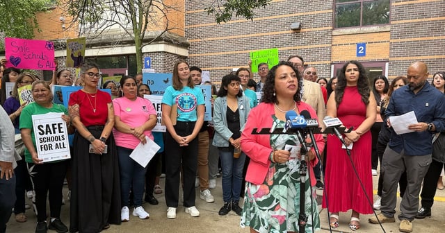 A photograph of a group of people holding signs standing behind a woman speaking behind a group of microphones all in front of a brick school building.