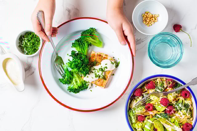 Children&#39;s hands eating fish with creamy sauce and broccoli in white plate, top view.