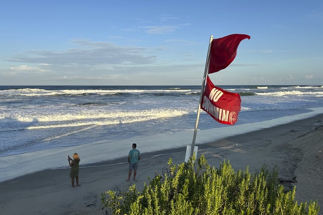 As waves from Hurricane Erin crash and “No Swimming” flags flap in the breeze, people walk on the beach in Nags Head, N.C., on Tuesday, Aug. 19, 2025. (AP Photo/Allen G. Breed)