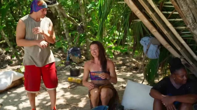 Three people in a tropical jungle; one is sitting on a box