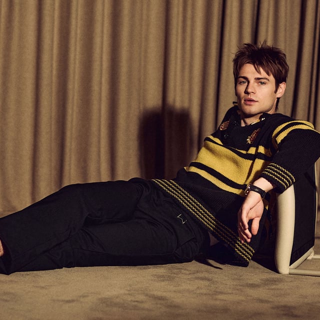 My Life With the Walter Boys actor Noah LaLonde for Who What Wear. He is sitting on the ground in front of a beige curtain leaning against a chair that is tipped on its side. He is wearing a yellow and black sweater with black pants and shoes.