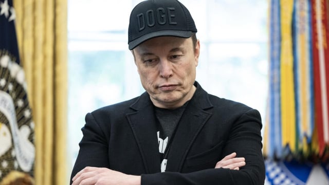Elon Musk, wearing a baseball cap with the word DOGE, stands with while crossing his arms.