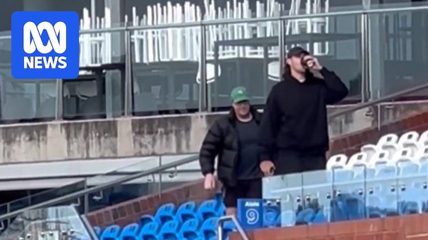 Sam Naismith pictured drinking a coffee at Whitten Oval, wears all black, another man behind wears black clothes and green cap. 