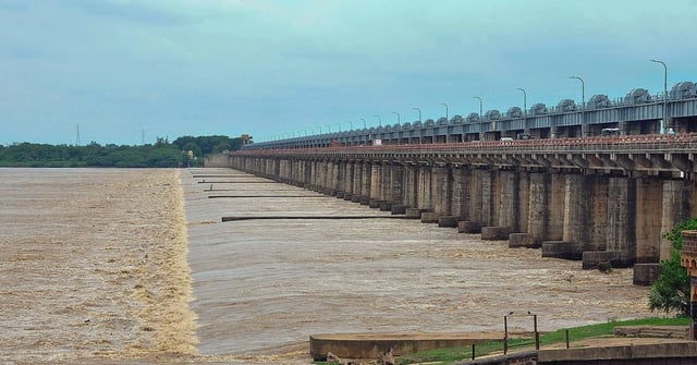 A wide view of Dowleswaram Barrage on the Godavari River, showing strong water flow beneath the long bridge-like structure with multiple pillars and gates. The cloudy sky and greenery in the background add to the scene, while vehicles can be seen moving on the road atop the barrage.
